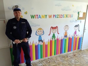 policjant stoi pod ścianą w przedszkolu, na ścianie widnieje napis witamy w przedszkolu