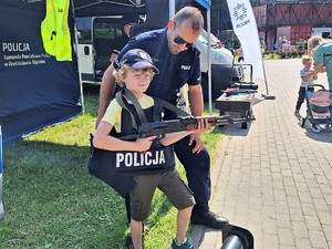 Na zdjęciu widać policjanta oraz pozującego do zdjęcia chłopca w kamizelce kuloodpornej i bronią