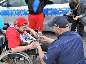 Na zdjęciu widać policjanta wraz z osobą siedzącą na wózku inwalidzkim. Policjant pokazuje tej osobie kajdanki