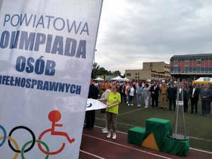 Na zdjęciu widnieje szyld z napisem Powiatowa Olimpiada Osób Niepełnosprawnych,a w tle zaproszeni goście